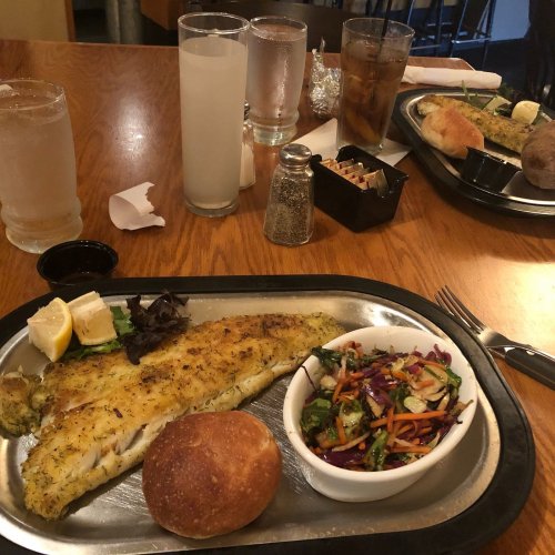A plated meal featuring baked fish, a side salad, a roll, and drinks on a wooden table in a restaurant setting.
