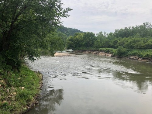 A calm river flows through a lush green landscape, surrounded by trees and gentle hills under a cloudy sky.