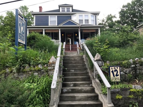 A blue house with a porch, surrounded by greenery, with a sign reading "Lot 12 Public Park" at the entrance.