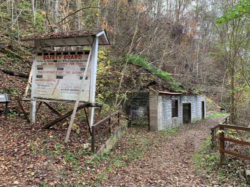 A weathered safety board stands beside a path leading to two small, abandoned buildings in a wooded area.