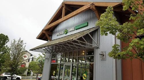 Exterior of a building with a metal facade and a sign that reads "Pickle Time," surrounded by trees and a cloudy sky.