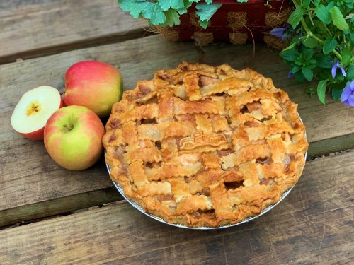 A freshly baked apple pie with a lattice crust, surrounded by two red apples on a wooden surface.