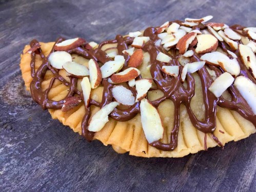 A golden pastry filled with chocolate and topped with sliced almonds, resting on a wooden surface.