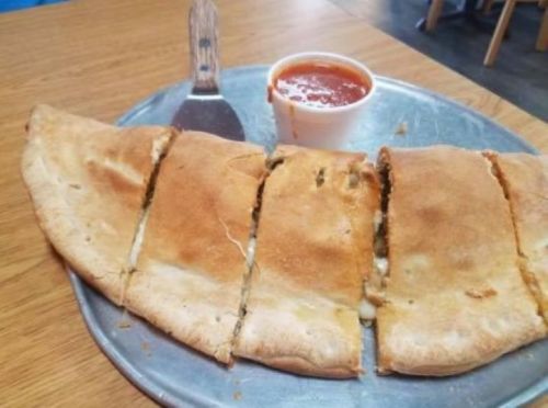A golden-brown calzone sliced into pieces, served with a small cup of marinara sauce on a metal plate.