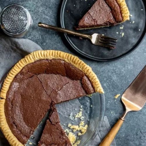 A chocolate pie with a slice removed, served on a black plate with a fork, on a textured surface.