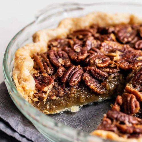 A close-up of a pecan pie in a glass dish, showcasing a golden crust and a rich, nutty filling topped with pecans.