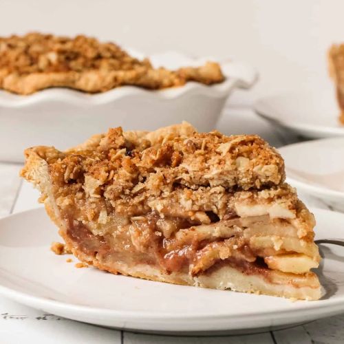 A slice of apple pie with a crumbly topping, served on a white plate, with a whole pie in the background.