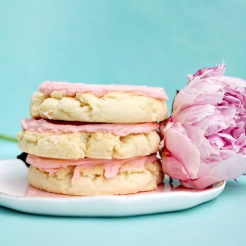 Three pink-frosted cookies stacked on a plate, accompanied by a blooming peony flower, against a light blue background.