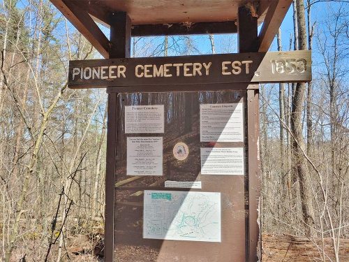 Sign for Pioneer Cemetery established in 1859, surrounded by trees, with informational posters and a map.