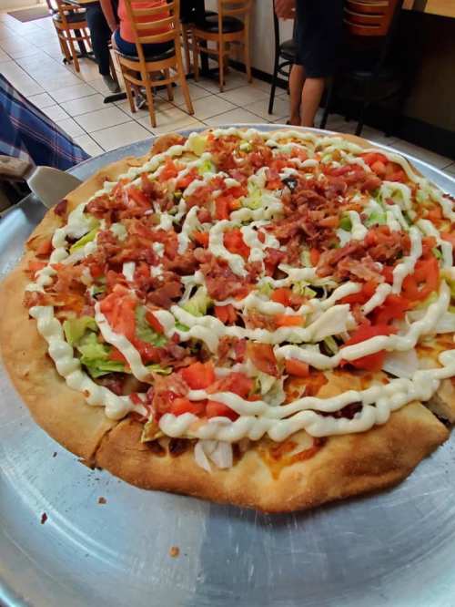 A large pizza topped with lettuce, tomatoes, bacon, and drizzled with mayonnaise, served on a metal tray.