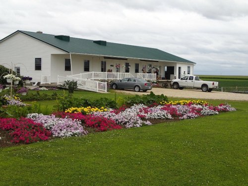 A white house with a green roof, surrounded by colorful flower beds and parked vehicles on a cloudy day.
