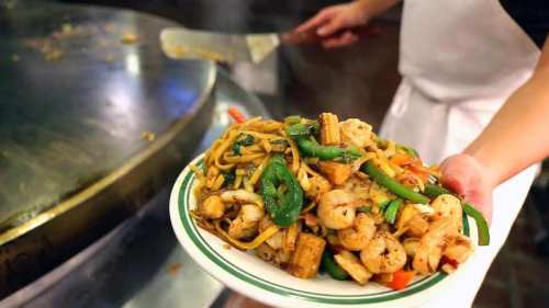 A chef holds a plate of stir-fried noodles with shrimp and vegetables, with a large cooking surface in the background.