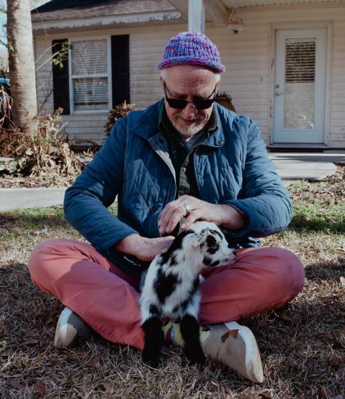 A man in a blue jacket and pink pants sits on the grass, gently petting a small black and white goat.