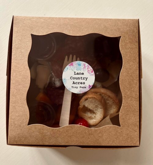 A brown takeout box with a clear window, labeled "Lane Country Acres Tiny Farm," containing assorted food items.
