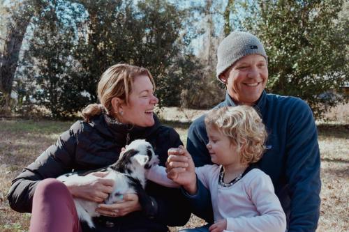 A woman, a man, and a young girl sit together outdoors, smiling and holding a small puppy in a sunny, green setting.