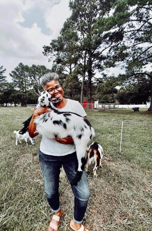 A smiling person holds a spotted goat in a grassy field, surrounded by trees and other goats in the background.