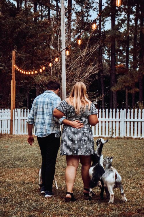 A couple walks hand in hand with two goats in a backyard, string lights glowing above in a wooded setting.