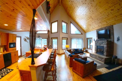 Cozy cabin interior featuring a wooden ceiling, stone fireplace, and open kitchen with bar seating. Bright natural light.