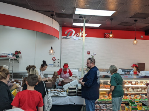 A busy deli with customers and staff, featuring a counter with food items and a sign that reads "Deli."