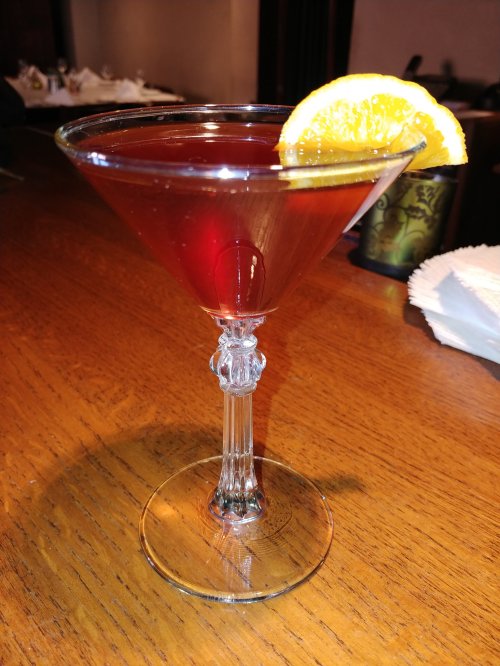 A vibrant red cocktail in a clear martini glass, garnished with a slice of orange, on a wooden table.