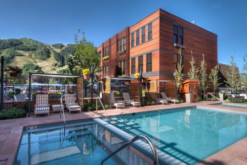 A modern building beside a clear pool, surrounded by lounge chairs and lush greenery, with mountains in the background.