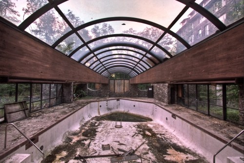 Abandoned indoor pool with a glass roof, surrounded by overgrown vegetation and debris on the floor.