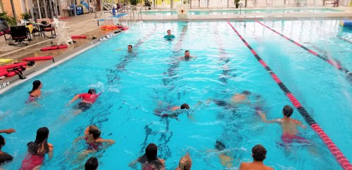 A group of people swimming in a large indoor pool with lanes marked by red and black lines. Lifeguard chairs are visible.
