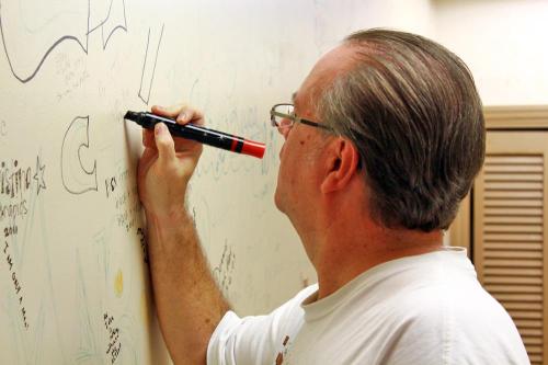 A man with glasses writes on a wall with a black marker, surrounded by various drawings and scribbles.