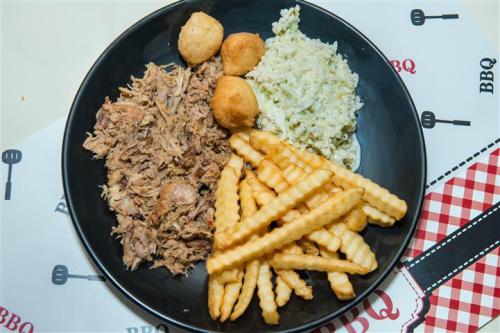 A black plate with pulled pork, rice, hush puppies, and crinkle-cut fries on a checkered tablecloth.