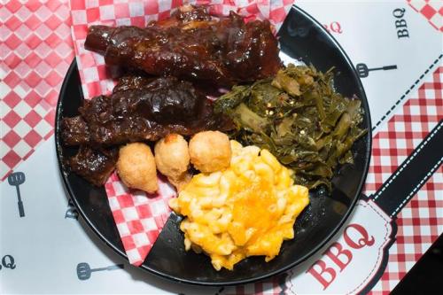 A plate of BBQ ribs, macaroni and cheese, collard greens, and cornbread balls on a checkered tablecloth.