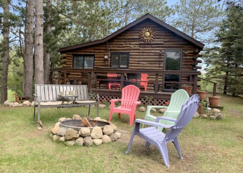 A cozy log cabin surrounded by trees, featuring a fire pit and colorful chairs in a serene outdoor setting.