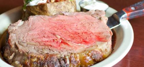 A close-up of a juicy slice of prime rib on a plate, with a baked potato and dipping sauce in the background.