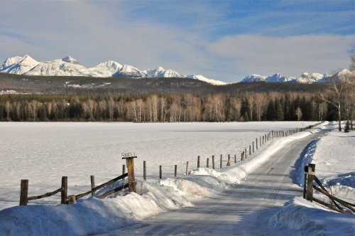 A snowy path leads to a frozen lake, with snow-capped mountains and trees in the background under a clear blue sky.