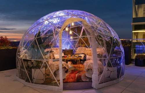 A cozy, illuminated geodesic dome on a rooftop, featuring plush seating and a warm atmosphere at dusk.