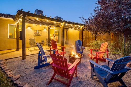 Cozy outdoor space with colorful chairs around a fire pit, illuminated by string lights, and a warm, inviting home in the background.