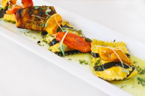 A gourmet dish featuring colorful ravioli topped with grilled seafood and garnished with microgreens on a white plate.