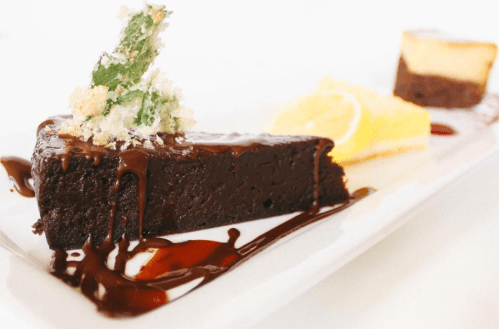 A slice of rich chocolate cake drizzled with chocolate sauce, garnished with mint and served with lemon and cheesecake.