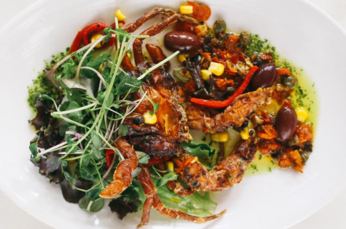 A plate featuring a cooked crab with vegetables, olives, and greens, garnished with herbs and colorful ingredients.