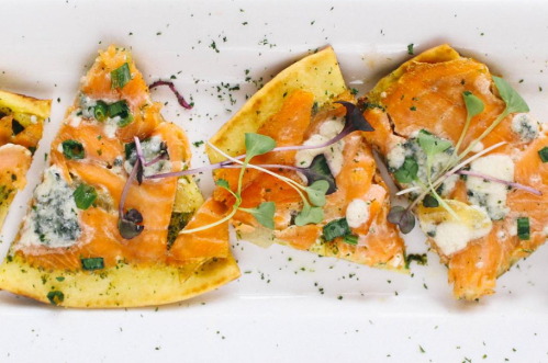 A plate of sliced flatbread topped with smoked salmon, blue cheese, herbs, and microgreens.