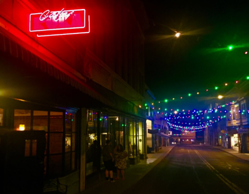 A dimly lit street at night, adorned with colorful string lights and a neon sign, creating a festive atmosphere.