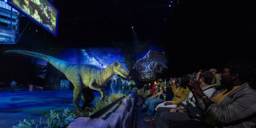 A lifelike dinosaur on stage with an audience watching, surrounded by a scenic backdrop and screens displaying images.