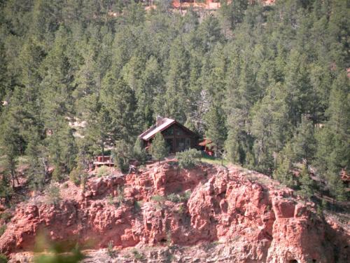 A rustic cabin perched on a rocky cliff, surrounded by dense green pine trees and red rock formations.