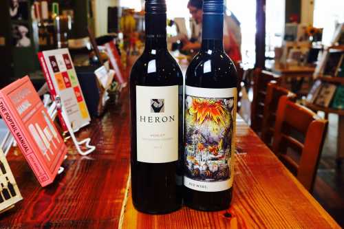 Two bottles of red wine on a wooden table, one labeled "Heron" and the other with a colorful, artistic design.