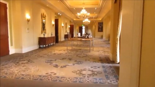 A spacious, elegantly decorated hallway with chandeliers, ornate rugs, and wooden furniture.