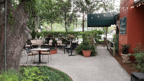 Outdoor seating area with tables and chairs, surrounded by greenery and string lights, leading to a covered entrance.