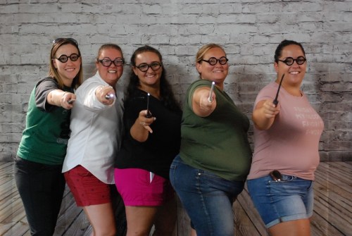 Five women in glasses pose playfully with wands against a brick wall backdrop, smiling and pointing forward.
