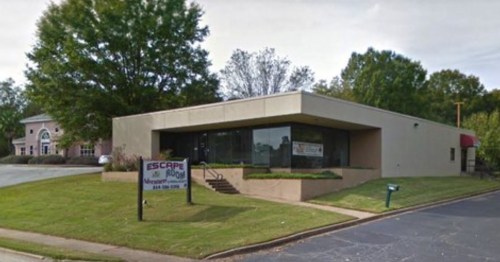 A single-story building with large windows and a sign for "Escape Room" in a grassy area. Trees are visible in the background.