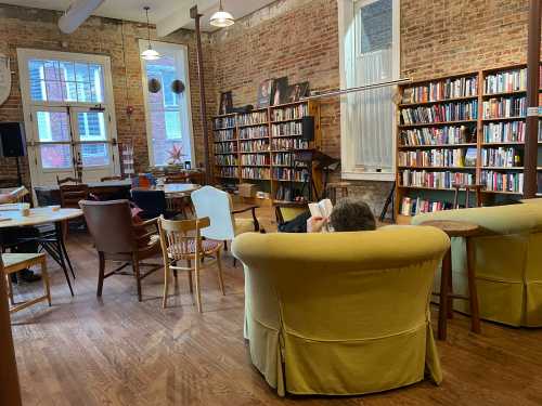 Cozy library with brick walls, bookshelves, and seating areas; a person reads in a yellow armchair.