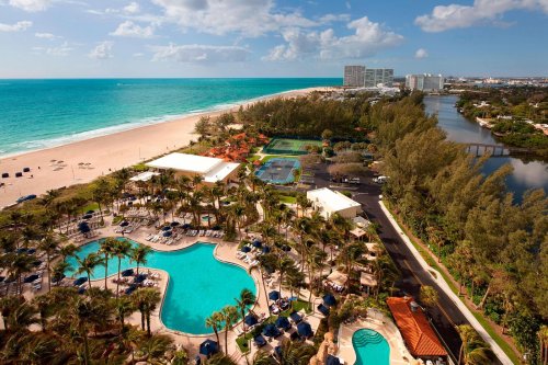 Aerial view of a resort with pools, palm trees, and a beach, overlooking turquoise waters and a clear blue sky.