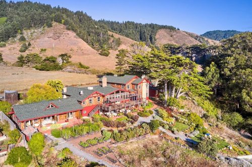Aerial view of a large, red-roofed house surrounded by gardens and hills, with trees and a clear blue sky.